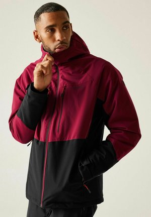 Mężczyzna w bordowo-czarnej kurtce z kapturem, zapinający na zamek kołnierz, z jedną ręką w kieszeni kurtki, patrzący w bok.