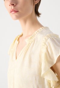 Lys gul bluse med V-hals, volangkrage og korte fladderermser. Har et teksturert stoff med et subtilt blomstermønster.