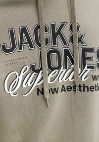 Φούτερ με κουκούλα σε ελιά από μείγμα βαμβακιού, με μεγάλο τυπωμένο κείμενο "JACK & JONES Superior" σε μαύρο και λευκό, με κορδόνια.