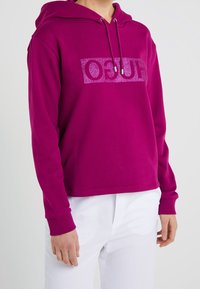 Sweat à capuche fuchsia en coton doux avec un logo pailleté sur le devant. Il comprend une capuche à cordon de serrage et des poignets côtelés, porté avec un pantalon blanc.