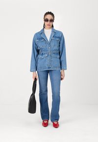 Denimjack met twee borstzakken en een ceintuur in de taille, gecombineerd met uitlopende jeans en rode schoenen. Geaccessoriseerd met zwarte zonnebril en een tas.