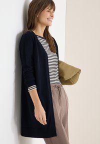 Femme souriante appuyée contre un mur, portant un cardigan bleu marine, une chemise rayée, un pantalon beige, tenant une veste olive pliée sous le bras.