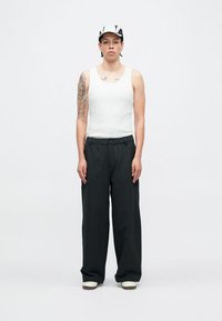 Balts topiņš, melnas plašas bikšu bikses un rakstains cepure. Uz kreisās rokas redzama ievērojama tetovējums, stāv uz vienkārša fona.