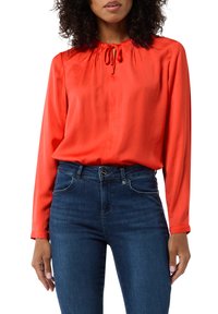 Czerwona satynowa bluzka z długimi rękawami i ściągaczem przy dekolcie, w zestawieniu z ciemnoniebieskimi dżinsami. Gładka tekstura i luźny krój.