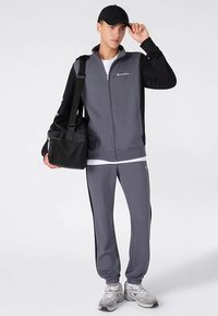 Gray zip-up jacket with black sleeves, paired with matching gray sweatpants. Model holds a black bag and wears a black cap and sneakers.