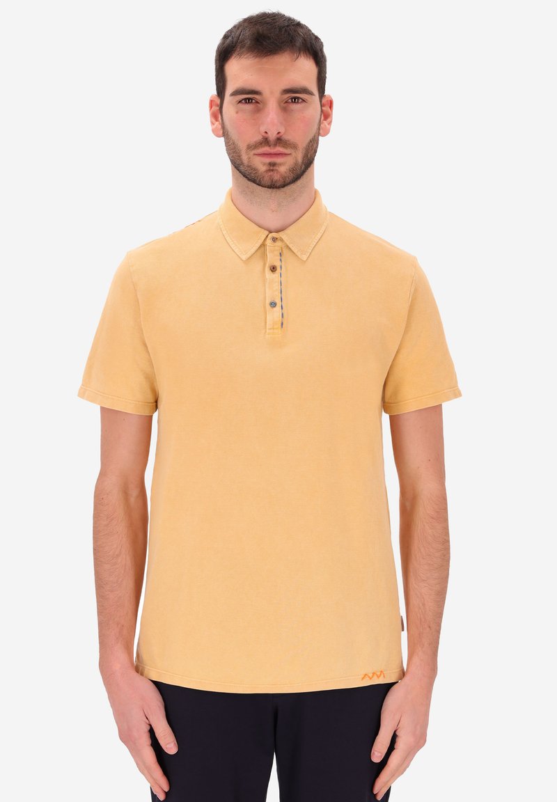 Uomo con capelli scuri e corti e barba, indossa una polo gialla a maniche corte e pantaloni neri, in piedi su uno sfondo chiaro e uniforme.