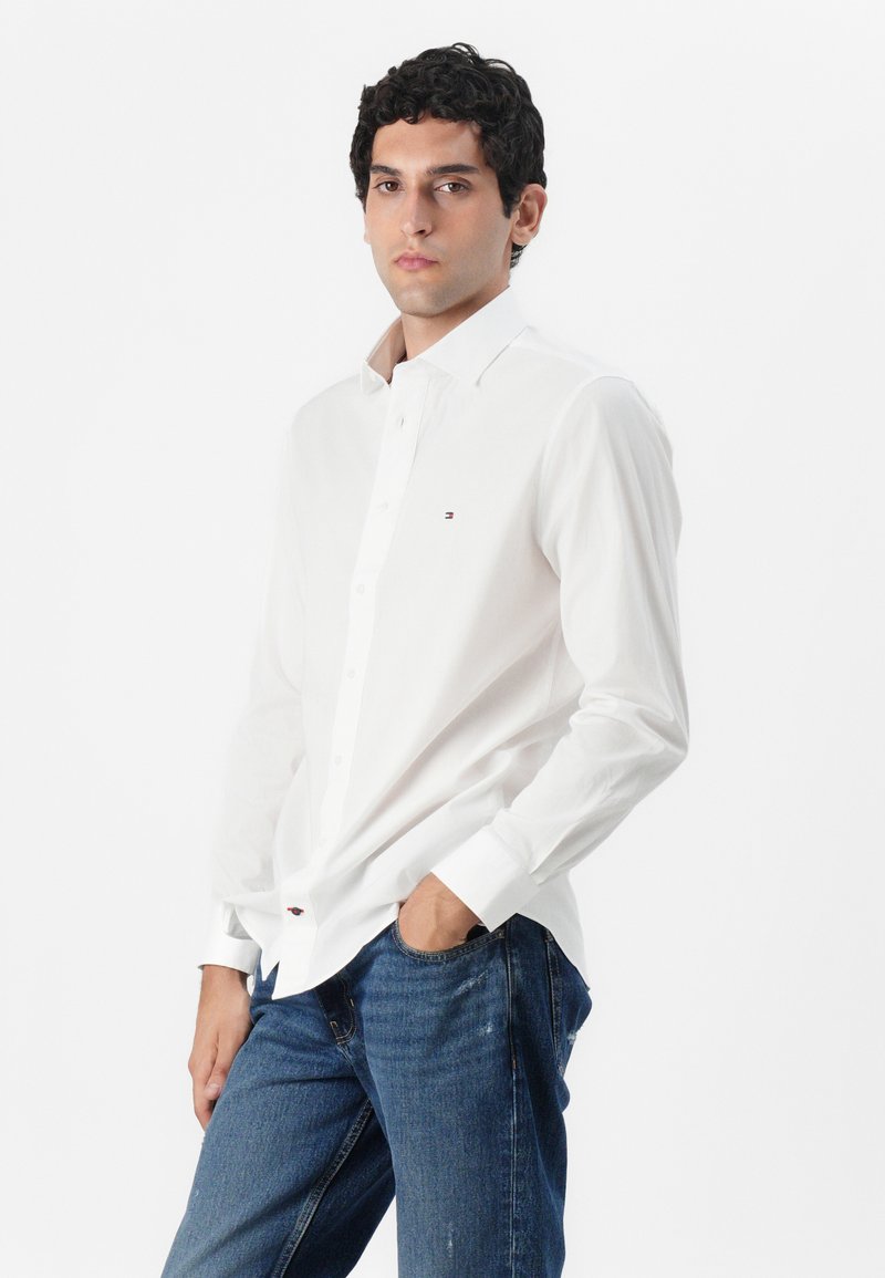 Chemise blanche à boutons avec un col classique, des manches longues et un logo brodé sur la poitrine gauche, associée à un jean bleu.