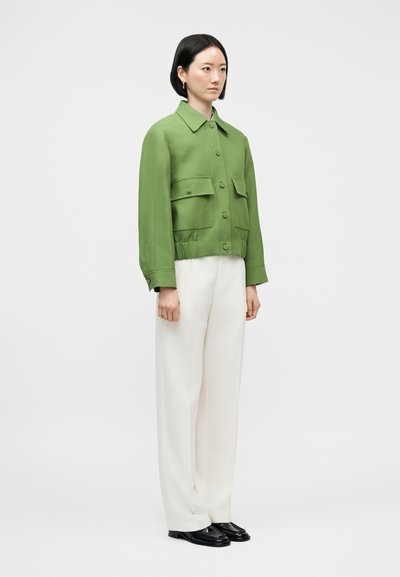 Femme portant une veste verte à boutons avec de grandes poches, un pantalon blanc à jambes larges et des chaussures noires, debout sur un fond clair uni.