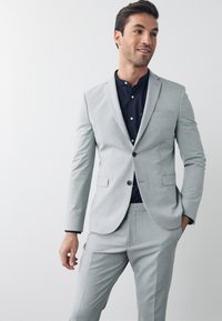 Completo grigio chiaro con giacca strutturata e pantaloni abbinati. Camicia blu scuro con piccoli pois bianchi. Chiusura con un solo bottone e tasche frontali.