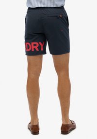 Mann trägt marineblaue Badeshorts mit rotem "DRY"-Schriftzug am linken Bein und braune Lederloafers, steht vor weißem Hintergrund.