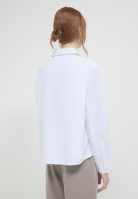 Camicia bianca a maniche lunghe con colletto e polsini con bottoni, caratterizzata da una trama liscia e una vestibilità comoda. La vista posteriore mostra dettagli di cucitura.