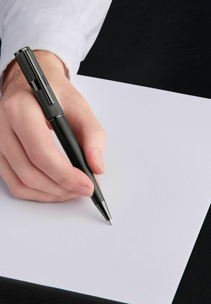 Czarny długopis trzymany nad pustą kartką białego papieru. Długopis posiada teksturowany uchwyt oraz spiczasty koniec, charakteryzuje się smukłym designem.