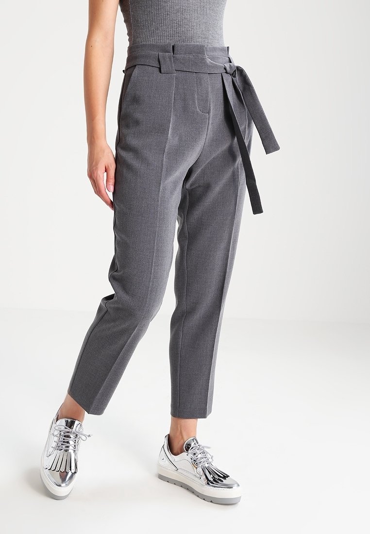 Kvinna i högmidjade grå avsmalnande byxor med tygbälte knutet på sidan och metalliska silverfärgade plattformsskor med snörning.