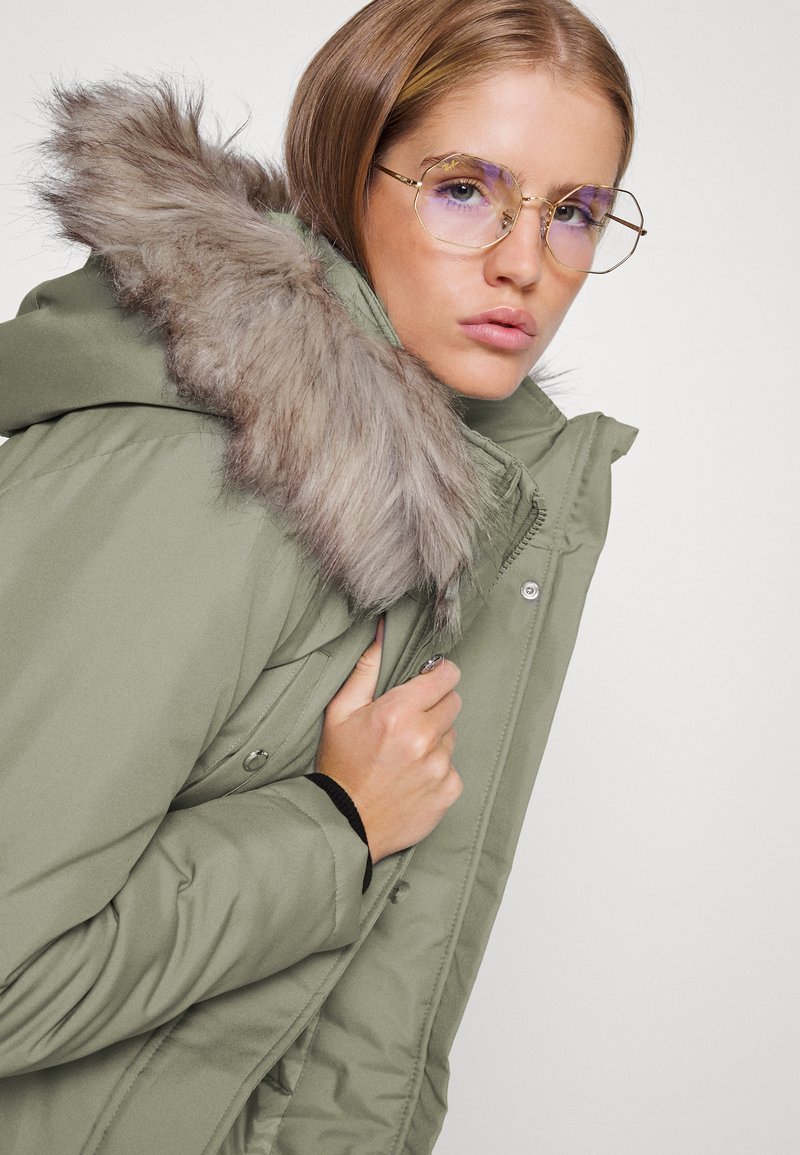 Doudoune verte olive avec un bord de capuche en fausse fourrure, fermeture éclair et poches latérales. Le modèle porte des lunettes rondes avec une teinte violette claire.