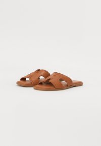 Brune lærsandaler med flad sål, prydet med udskæringer og syede kanter. Blød tekstur, afslappet design, åbentået stil.