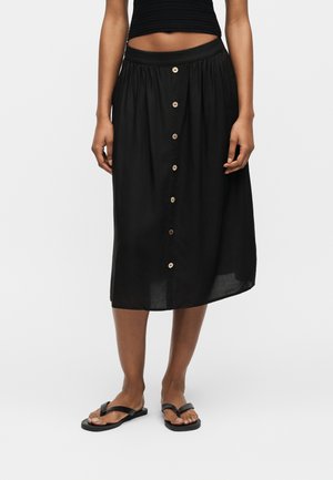 Jupe midi noire à boutons sur le devant, portée avec un crop top noir et des sandales noires type tongs, jambes et ventre visibles sur fond blanc.