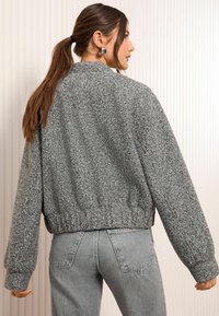 Grijze, gestructureerde bomberjack met een aansluitende tailleband en oversized mouwen, gecombineerd met lichtgrijze jeans. Gladde achtergrond met verticale lijnen.