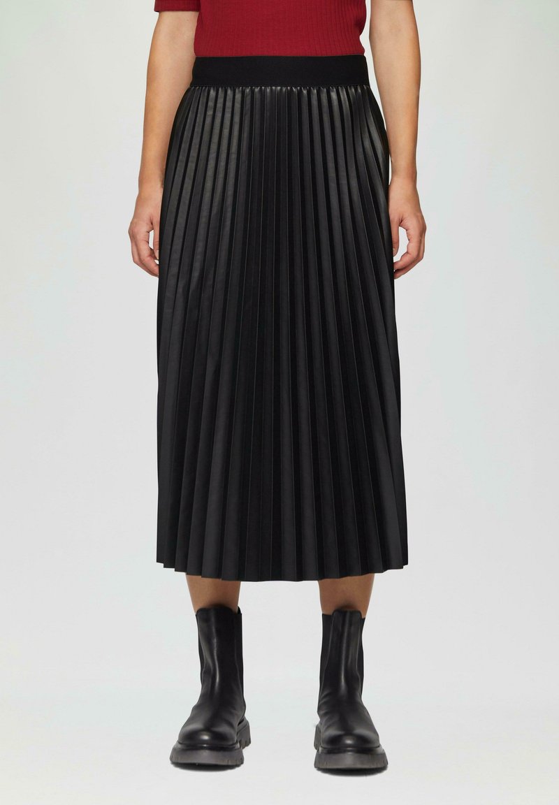 Jupe plissée noire en cuir, dotée d'une large taille et d'une silhouette fluide, associée à des bottes noires à cheville.