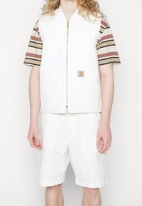 Weißer Canvas-Weste mit zwei Fronttaschen, goldener Reißverschluss und Carhartt-Logo; getragen über einem gestreiften Kurzarmhemd und weißen Shorts.