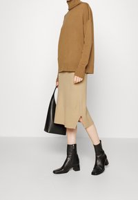 Beige polotröja med ribbade manschetter, kombinerad med en beige midikjol med en sidoöppning. Svarta läderstövletter och en svart väska.