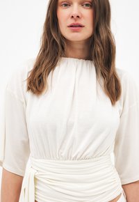 Mujer con cabello castaño claro, vestida con una blusa blanca fruncida, de mangas cortas y con un lazo ajustado en la cintura, sobre un fondo liso.