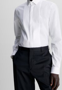 Witte overhemden met een geplisseerde voorkant, lange mouwen en button-down kraag, gecombineerd met zwarte formele broek. Gladde katoenstof.