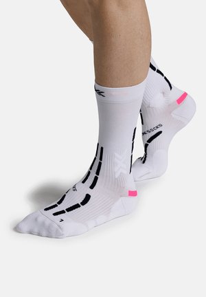 Benen met witte sportsokken met zwarte strepen en roze accenten, staand op een witte oppervlakte, met het ontwerp en de pasvorm van de sokken zichtbaar.