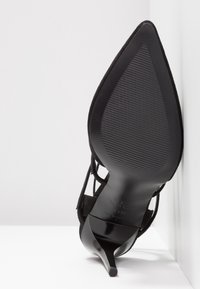 Chaussure à talon aiguille noir à bout pointu vue de la semelle, positionnée contre un mur blanc et une surface blanche.