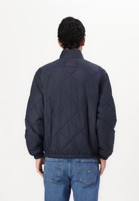 Mann in einer gesteppte marineblauen Jacke und blauen Jeans, der mit dem Rücken zur Kamera vor einem schlichten weißen Hintergrund steht.