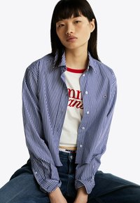 Camisa de botones a rayas azul y blanca con mangas largas, combinada con una camiseta blanca con letras rojas. Jeans de mezclilla visibles.
