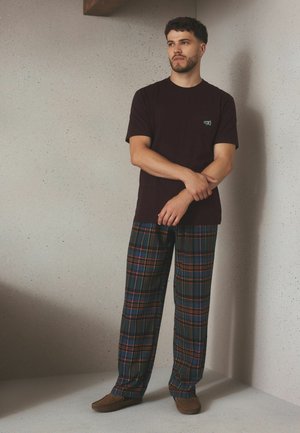 Mężczyzna z krótkimi kręconymi włosami, ubrany w ciemny t-shirt, niebiesko-pomarańczowe spodnie w kratę oraz brązowe kapcie, stojący przy jasnej cętkowanej ścianie.
