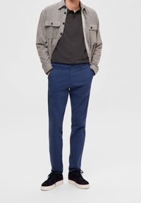 Šedá košile na knoflíky přes tmavé polo tričko; modré chino kalhoty kombinované s tmavými teniskami. Oblečení ukazuje ležérní, vrstvený vzhled.