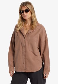 Bruine button-up shirt met een relaxte pasvorm, twee borstzakken en een zachte, gestructureerde stof. Draagt bij donkere broek en met een gemusterd zonnebril.