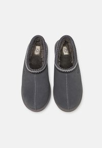 Pantofole in suede grigio con una morbida pelliccia interna, apertura elasticizzata e cuciture a contrasto. Presentano il marchio UGG sul lato.