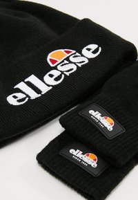 Černá pletená čepice a sada rukavic bez prstů s bílým logem "ellesse" a nášivkou s barevným emblémem; měkká textura a žebrovaný design.