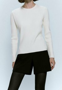 Biała, prążkowana bluzka z długim rękawem połączona z czarnymi szortami o wysokim stanie, noszona z czarnymi rajstopami. Ubranie charakteryzuje się prostym, zorganizowanym krojem.