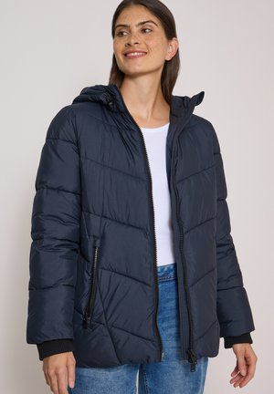 Femme portant une doudoune bleu marine avec une capuche, une chemise blanche et un jean bleu, souriante et regardant légèrement sur le côté.