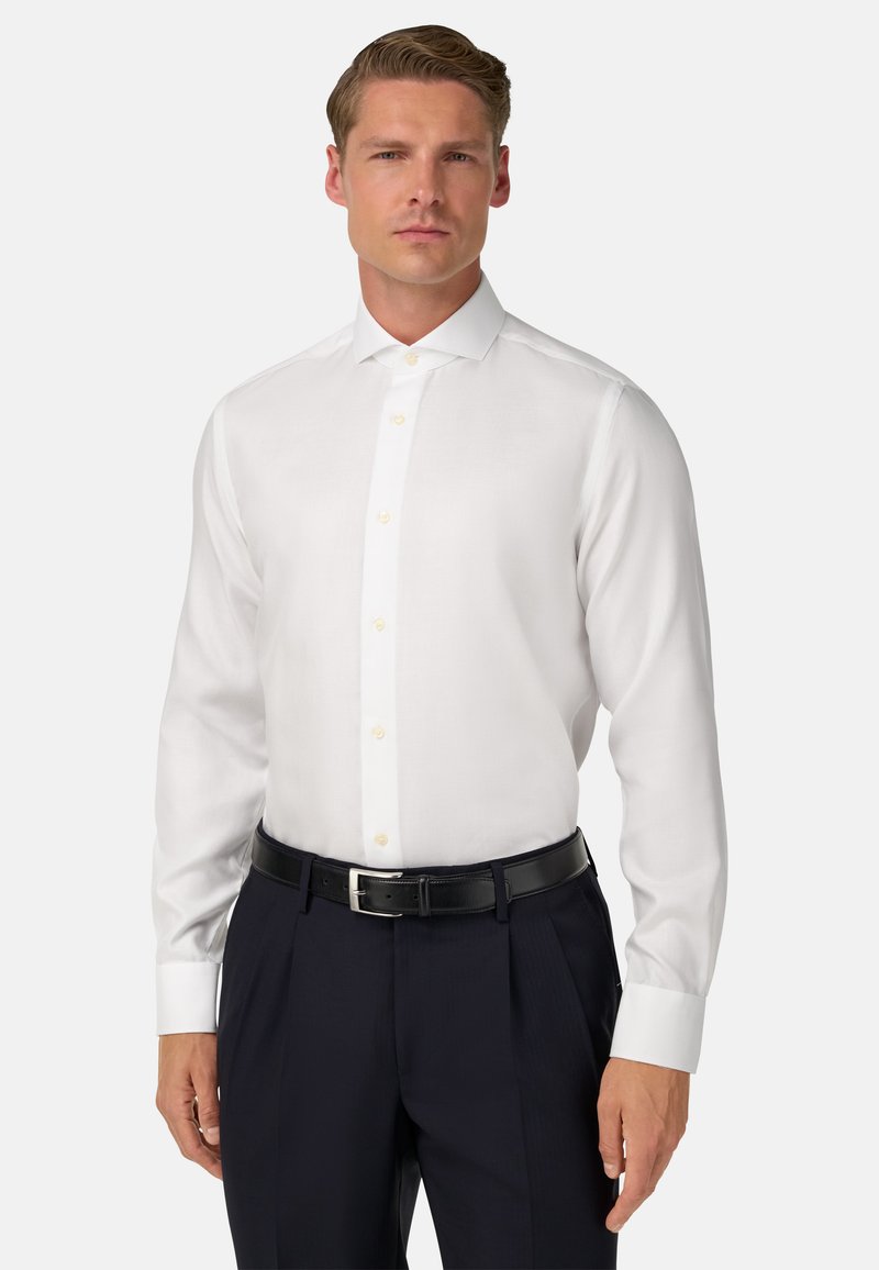 Bílá dlouhá košile s knoflíky vyrobená z hladké látky, s klasickým límečkem a žlutými knoflíky, v kombinaci s tmavými kalhotami a opaskem.