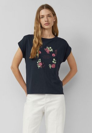 Kobieta z długimi blond włosami, ubrana w granatowy t-shirt z różowymi i zielonymi kwiatowymi wzorami oraz białe spodnie, stojąca na jednolitym tle.
