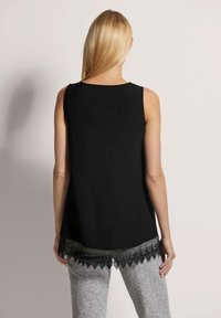 Schwarzes ärmelloses Oberteil mit abgerundetem Saum und Spitzenbesatz am unteren Rand. Graue gemusterte Hose sichtbar, die ein mehrschichtiges Design betont.