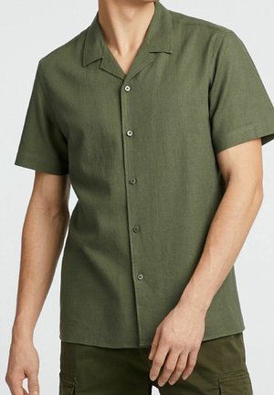 Homme portant une chemise boutonnée olive à manches courtes avec col camp et un pantalon cargo assorti, debout devant un fond uni.