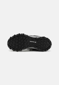 Semelle en caoutchouc noir texturé avec des motifs de crampons hexagonaux et circulaires bien visibles. Présente un logo imprimé près du talon pour une meilleure adhérence et stabilité.