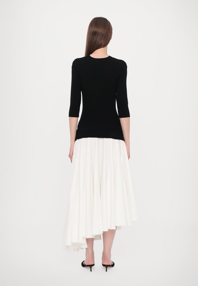 Schwarzes, figurbetontes Strickoberteil mit drei Viertelärmeln, kombiniert mit einem fließenden weißen asymmetrischen Rock. Das Outfit hat eine strukturierte Oberfläche und eine elegante Silhouette.