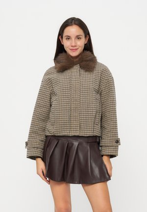 Jeune femme portant une veste à carreaux avec un col en fourrure marron et une jupe plissée en cuir brun foncé, debout et souriante.