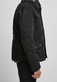 Muž oblečený v černé zapínací bundě a černých kalhotách, stojící bokem na jednoduchém světle šedém pozadí.