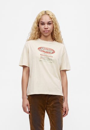 Giovane con lunghi capelli biondi ricci che indossa una maglietta beige Replay con grafica e pantaloni marroni, in piedi su uno sfondo bianco.