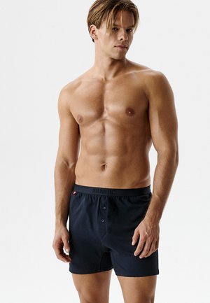 Młody mężczyzna bez koszulki, o krótkich włosach, ubrany w granatowe bokserki z guzikiem i paskiem z napisem "ATLANTIC" na białym tle.