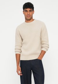 Maglione beige a maglia con un motivo testurizzato e polsini a costine, indossato con pantaloni blu scuro. Collo rotondo semplice.