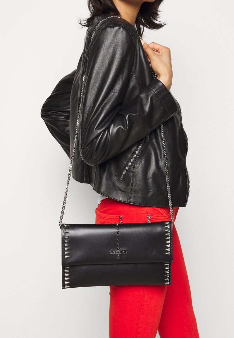 Femme portant une veste en cuir noire et un pantalon rouge tenant un sac bandoulière en cuir noir avec des clous argentés et une chaîne.