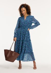 Pitkähiha, kerroksellinen mekko sinivihreällä paisley-kuviolla. Siinä on avainaukko-olkaintoppi ja hulmuava, puoliksi läpinäkyvä hame. Ruskea laukku ja nauhaliuskat.